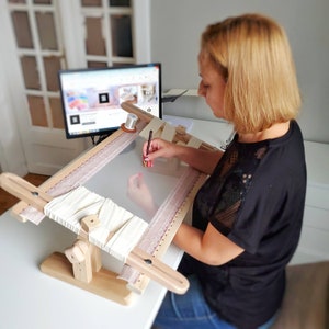 Op de afbeelding: Een persoon gebruikt een naald om te borduren op een witte stof die gespannen is op een houten borduurring. De ring is bevestigd aan een houten standaard. De persoon zit aan een tafel met een computermonitor op de achtergrond.