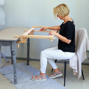Puede incluir: Una mujer se sienta en una mesa usando un soporte de aro de bordado de madera. El soporte está sujeto a la mesa con una abrazadera. La mujer lleva una camisa negra y pantalones blancos.