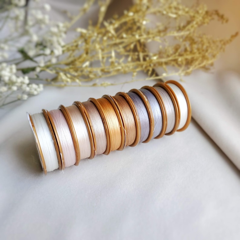 May include: A stack of spools of thread in various shades of white, beige, and gold. The spools are arranged in a graduated size order, with the largest spool at the bottom and the smallest spool at the top.