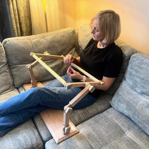 May include: A wooden embroidery stand with a rectangular base and adjustable arms. The stand is designed to hold a fabric frame, and is shown in use. The frame is made of light-colored wood and is positioned on a light-colored wooden base.