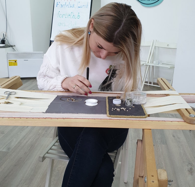 May include: A woman is seated at a table with a wooden frame, working on a needlepoint project. She is using a needle and thread to create a design on a piece of fabric stretched over a frame. There are beads and other supplies on the table.