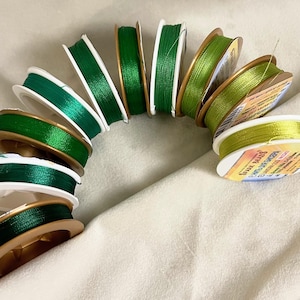 May include: A collection of spools of green and yellow thread, wound around white plastic bobbins with gold trim. The spools are arranged in a semi-circle on a white surface.