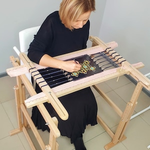 Könnte beinhalten: Eine Person sitzt an einem hölzernen Stickrahmen und arbeitet an einem farbenfrohen Stickprojekt. Der Rahmen hat verstellbare Beine und eine ebene Fläche zum Arbeiten. Das Stickprojekt zeigt ein geometrisches Muster mit blauen, gelben und grünen Fäden.