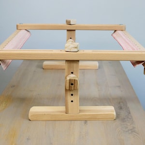 K&ouml;nnte beinhalten: Ein Holzst&auml;nder f&uuml;r einen Stickrahmen mit einem roten und wei&szlig;en Stoff, der &uuml;ber den Rahmen gespannt ist. Der St&auml;nder hat einen Sockel mit drei Beinen und einen vertikalen Pfosten mit einstellbarer H&ouml;he.