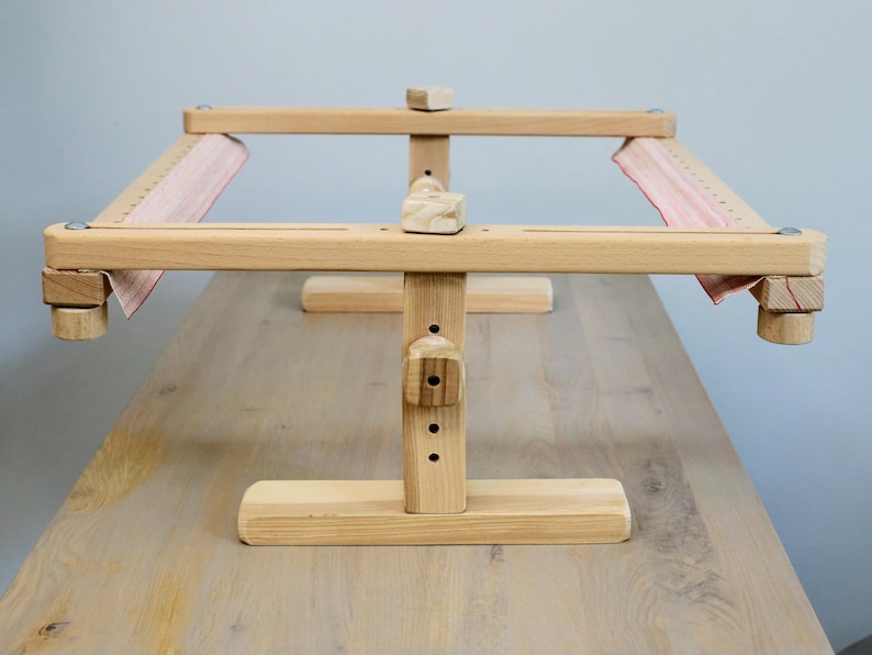 K&ouml;nnte beinhalten: Ein Holzst&auml;nder f&uuml;r einen Stickrahmen mit einem roten und wei&szlig;en Stoff, der &uuml;ber den Rahmen gespannt ist. Der St&auml;nder hat einen Sockel mit drei Beinen und einen vertikalen Pfosten mit einstellbarer H&ouml;he.