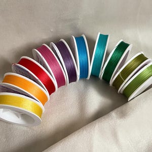 Op de afbeelding: Een set van tien spoelen borduurgaren in verschillende kleuren, waaronder geel, oranje, rood, roze, paars, blauw, groen en bruin. De spoelen zijn in een halve cirkel op een wit oppervlak gerangschikt.