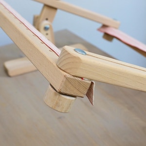 K&ouml;nnte beinhalten: Nahaufnahme eines Holzst&auml;nders f&uuml;r einen Stickrahmen mit einem roten und wei&szlig;en Stoffband. Der St&auml;nder ist aus Naturholz gefertigt und hat ein schlichtes Design.