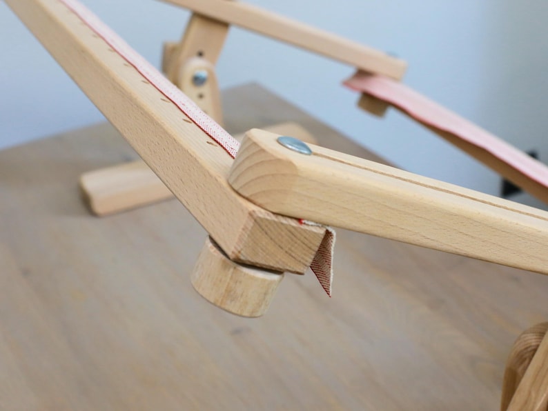 K&ouml;nnte beinhalten: Nahaufnahme eines Holzst&auml;nders f&uuml;r einen Stickrahmen mit einem roten und wei&szlig;en Stoffband. Der St&auml;nder ist aus Naturholz gefertigt und hat ein schlichtes Design.