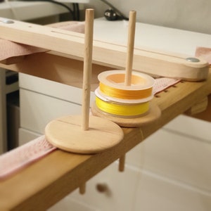 Peut inclure: Des bobines de fil jaune et orange sont placées sur des supports en bois. Les supports sont posés sur une table en bois avec un tissu rose clair.