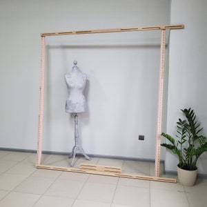 Op de afbeelding: Een houten frame met een lichte afwerking, ontworpen voor textielwerk. Het frame is rechthoekig, met gelijkmatig verdeelde gaten langs de zijkanten. Een paspop staat in het frame. Een potplant staat op de achtergrond.