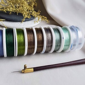 May include: A set of eight spools of elastic thread in various colours, including blue, green, brown, grey, and clear. The spools are arranged in a row, with the clear spool at the end. A wooden needle tool is also included.
