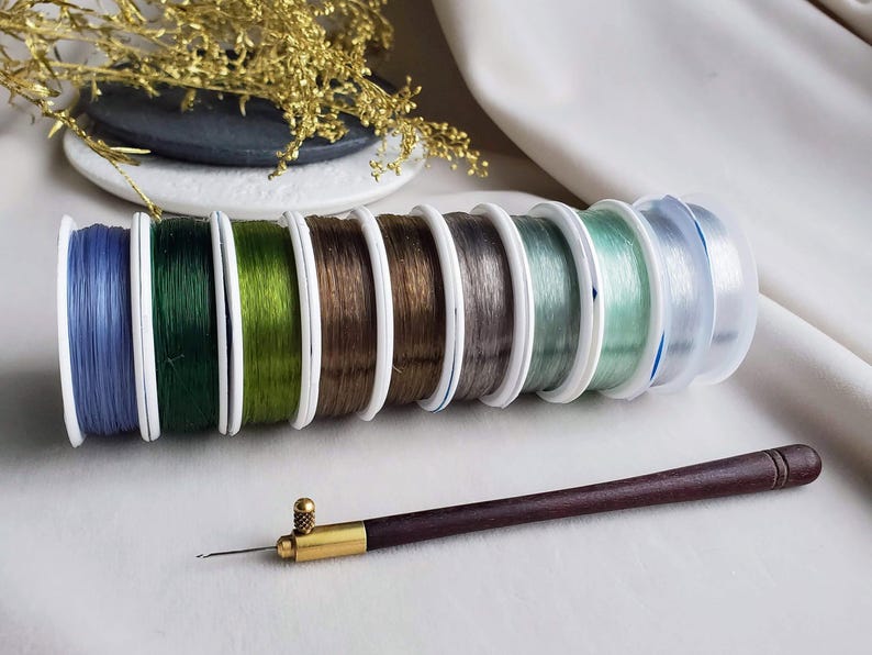 May include: A set of eight spools of elastic thread in various colours, including blue, green, brown, grey, and clear. The spools are arranged in a row, with the clear spool at the end. A wooden needle tool is also included.
