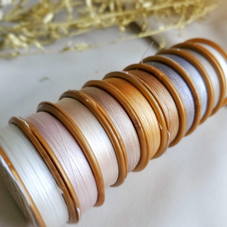 May include: A stack of spools of thread in various shades of white, beige, and gold. The spools are wound with delicate thread and have a wooden finish.