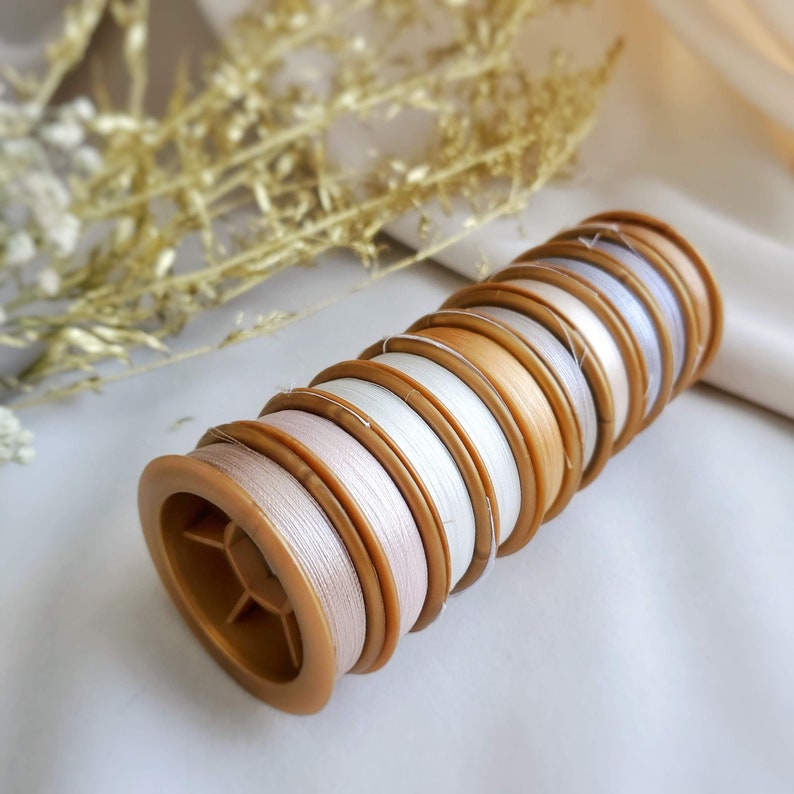 May include: A stack of spools of white and beige thread wound around brown plastic bobbins. The spools are arranged in a graduated size order, with the largest spool at the bottom and the smallest spool at the top.