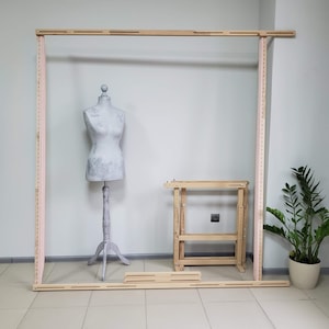 Puede incluir: Un marco de madera con un maniquí dentro, una pequeña mesa de madera con estantes y una planta en maceta. El marco está hecho de madera clara y tiene una serie de agujeros a los lados. El maniquí es gris y tiene un patrón floral en el torso. La mesa está hecha de madera clara y tiene dos estantes. La planta es verde y está en una maceta marrón.