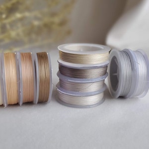 May include: Four spools of thread in different shades of beige and gray, wound around clear plastic spools. The spools are arranged in a row on a white surface.