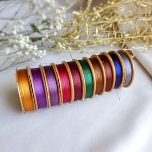Puede incluir: Una colección de carretes de hilo de varios colores, incluyendo dorado, morado, rojo, verde, marrón y azul. Los carretes están dispuestos en una pila, con el carrete dorado en la parte inferior y el carrete azul en la parte superior.