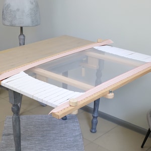 May include: A wooden embroidery hoop with a white fabric stretched across it. The hoop is attached to a wooden frame with a ruler and a knob on each end. The frame is sitting on a table.