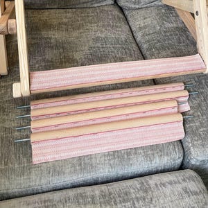 May include: A wooden embroidery frame stand with a light-colored wood finish and adjustable side supports. The frame includes a horizontal bar with a pink fabric strip attached, along with four additional wooden rollers featuring pink fabric strips.