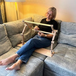 Può includere: Un supporto da ricamo in legno con una struttura chiara e una base rettangolare. Il supporto è regolabile e progettato per contenere il tessuto per lavori di cucito. Il supporto viene utilizzato su un divano grigio. Il supporto è lungo circa 50 cm.