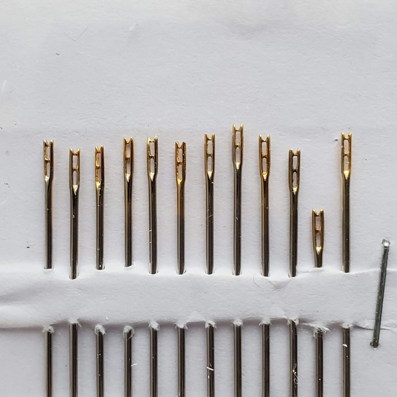 May include: A set of 12 gold-tipped needles in a white plastic case. The needles are arranged in rows and are all the same size.