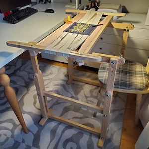 May include: A wooden embroidery stand with a beige canvas stretched across it. The stand has adjustable height and a built-in chair. The canvas is decorated with a colorful embroidery design.