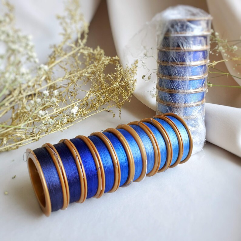 May include: A stack of blue and gold spools of thread. The spools are arranged in a graduated size, with the largest spool at the bottom and the smallest spool at the top. The spools are wrapped in blue thread and have gold trim.