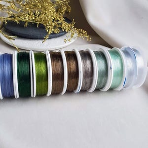 May include: Assortment of eight spools of elastic cord in various colours, including blue, green, brown, grey, and clear. The spools are arranged in a row, with the clear spool at the end.