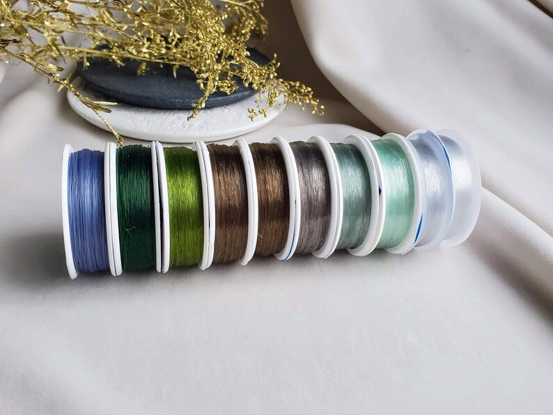 May include: Assortment of eight spools of elastic cord in various colours, including blue, green, brown, grey, and clear. The spools are arranged in a row, with the clear spool at the end.