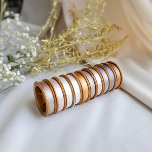 May include: A stack of spools of thread in various shades of white, beige, and grey. The spools are arranged in a circular pattern and are held together by a gold-coloured plastic ring.