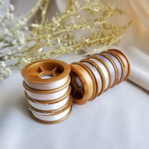 May include: A set of spools of thread in various shades of white and beige. The spools are wound with thread and are arranged in a stack. The spools are made of plastic and have a gold colour.