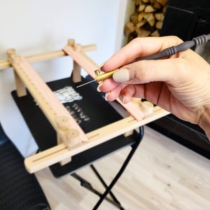 May include: A punch needle tool with a dark gray textured handle and a brass tip, held in position above a light-colored wooden embroidery frame. The frame features adjustable horizontal bars with circular wooden knobs and pink fabric strips.