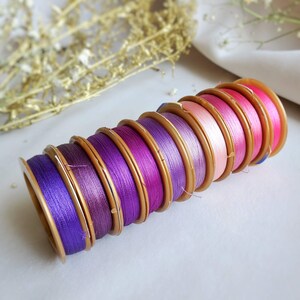 Puede incluir: Una pila de ocho carretes de hilo en varios tonos de morado y rosa. Los carretes están enrollados con hilo y tienen una carcasa de plástico dorada.