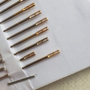 May include: A close-up of a set of gold-tipped needles for embroidery or sewing. The needles are arranged in a fan shape on a white surface.