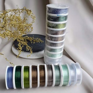 May include: A set of eight spools of clear elastic thread in various shades of green and blue. The spools are arranged in a row on a white surface.