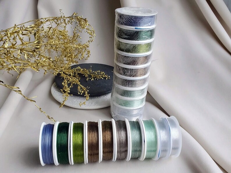 May include: A set of eight spools of clear elastic thread in various shades of green and blue. The spools are arranged in a row on a white surface.