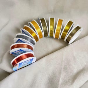 Op de afbeelding: Een verzameling spoelen met metallic draad in verschillende kleuren, waaronder goud, zilver, koper en rood. De spoelen zijn in een gebogen lijn gerangschikt, met de merknaam "ALLURE" zichtbaar op sommige labels. De draad is voor knutselen of sieraden maken.