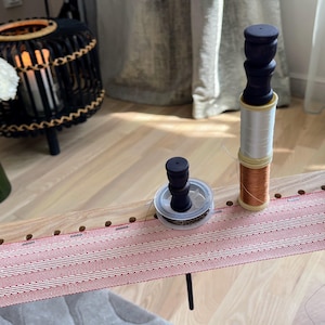 Op de afbeelding: Een houten weefgetouw met rood en wit geweven stof. Spoelen met wit en koperkleurig garen staan op het weefgetouw, samen met een kleinere spoel met donker garen. Een zwarte decoratieve lamp en bloemen staan op de achtergrond.