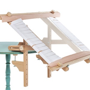 May include: A wooden embroidery hoop stand with a white fabric stretched across the hoop. The stand is attached to a table with a clamp.