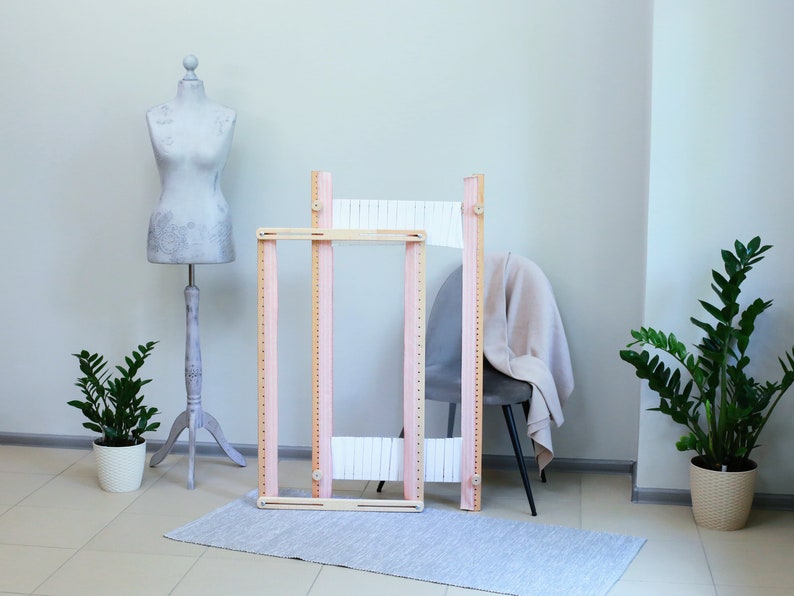 May include: Two wooden weaving looms with white string stretched across them. The looms are pink and natural wood and are standing on a light gray rug.
