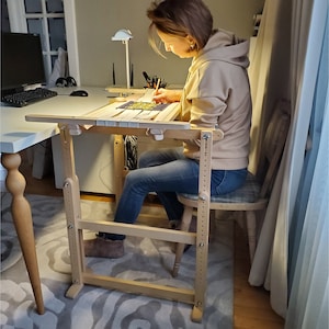 Può includere: Un cavalletto da ricamo in legno con altezza regolabile, con una persona seduta e che lavora a un progetto. Il cavalletto ha una finitura in legno chiaro ed è progettato per un'artigianato confortevole.