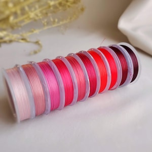May include: A set of eight spools of thread in various shades of pink and red. The spools are arranged in a gradient from light pink to dark red.