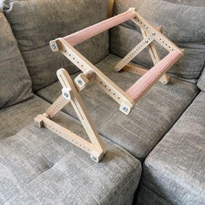 May include: A wooden embroidery hoop stand with a pink fabric insert. The stand is made of light-colored wood and features adjustable arms and a rectangular frame. It is designed to hold embroidery projects and is shown on a gray couch.