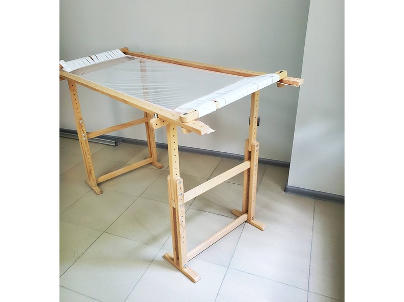May include: A wooden embroidery stand with a white canvas stretched across the frame. The stand is adjustable in height and has a simple, functional design.