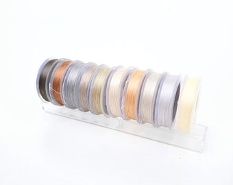 Può includere: Un set di dieci bobine di filo in varie tonalità di beige, marrone e bianco. Le bobine sono disposte in un porta-bobine di plastica trasparente.