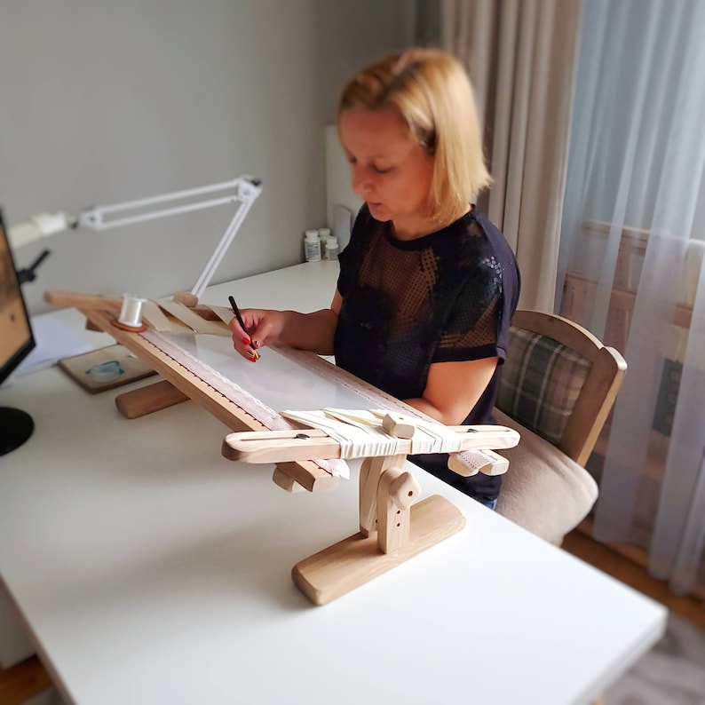 Op de afbeelding: Een houten borduurstandaard met een witte stof eroverheen gespannen. Een persoon gebruikt een potlood om de stof te markeren. De standaard staat op een witte tafel.