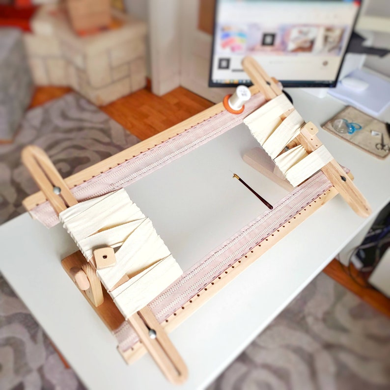 Op de afbeelding: Een houten borduurring met een witte stof eroverheen gespannen. De ring is gemonteerd op een standaard en een naald ligt op de stof.