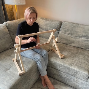 May include: A wooden embroidery hoop stand with adjustable height and angle. The stand is made of light-colored wood and features a rectangular frame to hold the fabric. The stand is designed to sit on a flat surface.