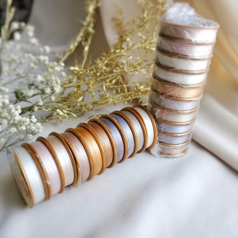 May include: A collection of spools of thread in various shades of white, beige, and gold. The spools are arranged in a stack, with the gold spools at the bottom and the white spools at the top.
