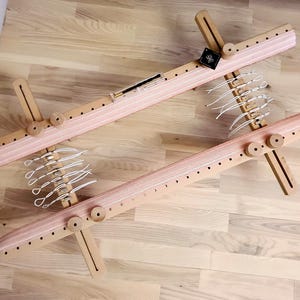 May include: A wooden loom with a pink and white warp thread. The loom has a series of pegs and a wooden frame.